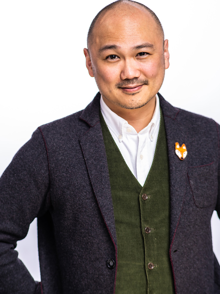 Headshot of Derek Kwan, an East Asian man in his 40s with a shaved head, and stubble moustache. He smiles at the camera with his hands on his hips. He is wearing a dark grey jacket, forest green corduroy vest, white shirt, and there is a small needle-felted head of a fox on his lapel