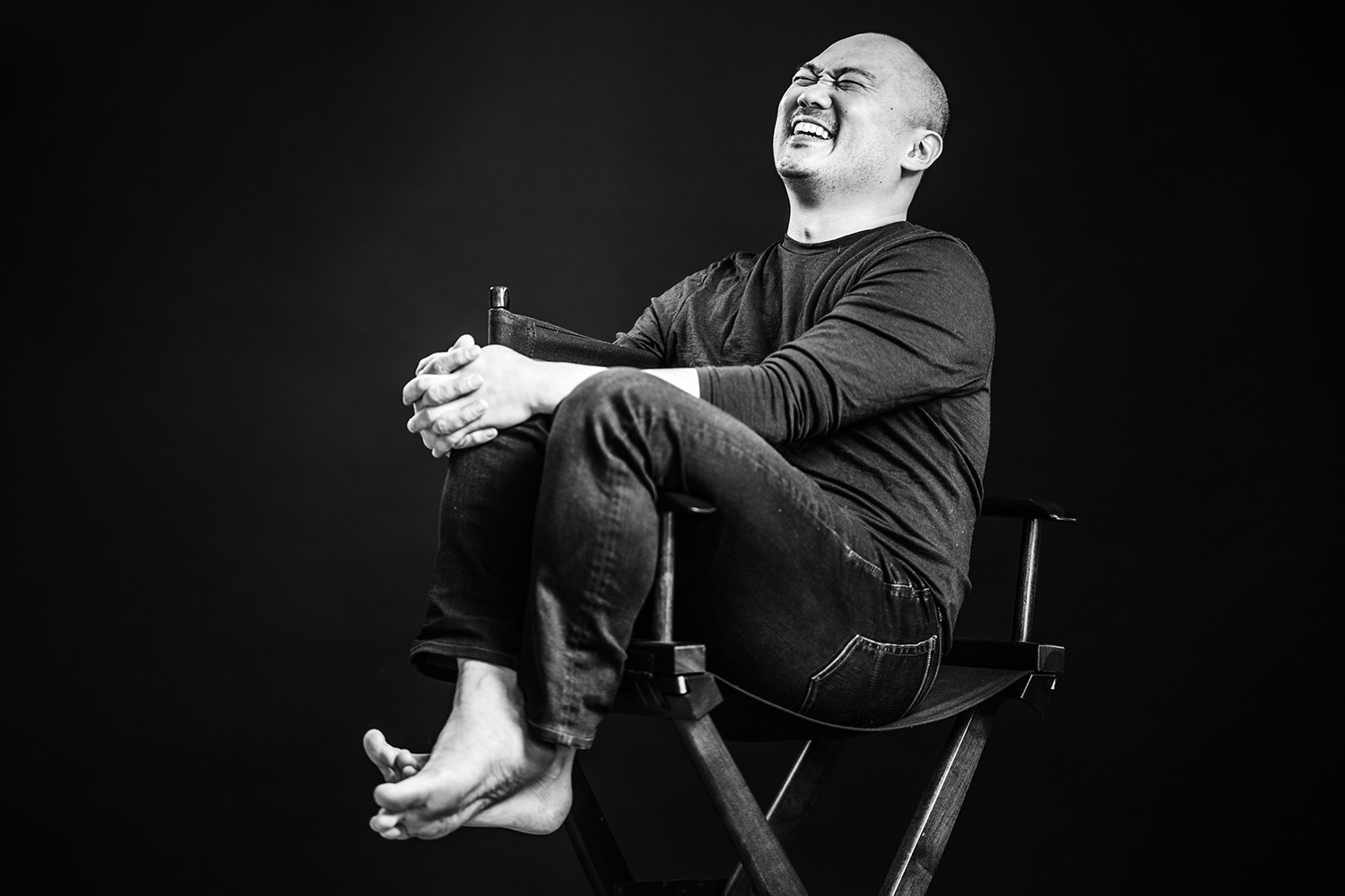 Black and white photo of Derek Kwan, sitting sideways in a high canvas chair with his knees draped over on of the arms of the chair. His hands clasp one knee and his head is bent back laughing.