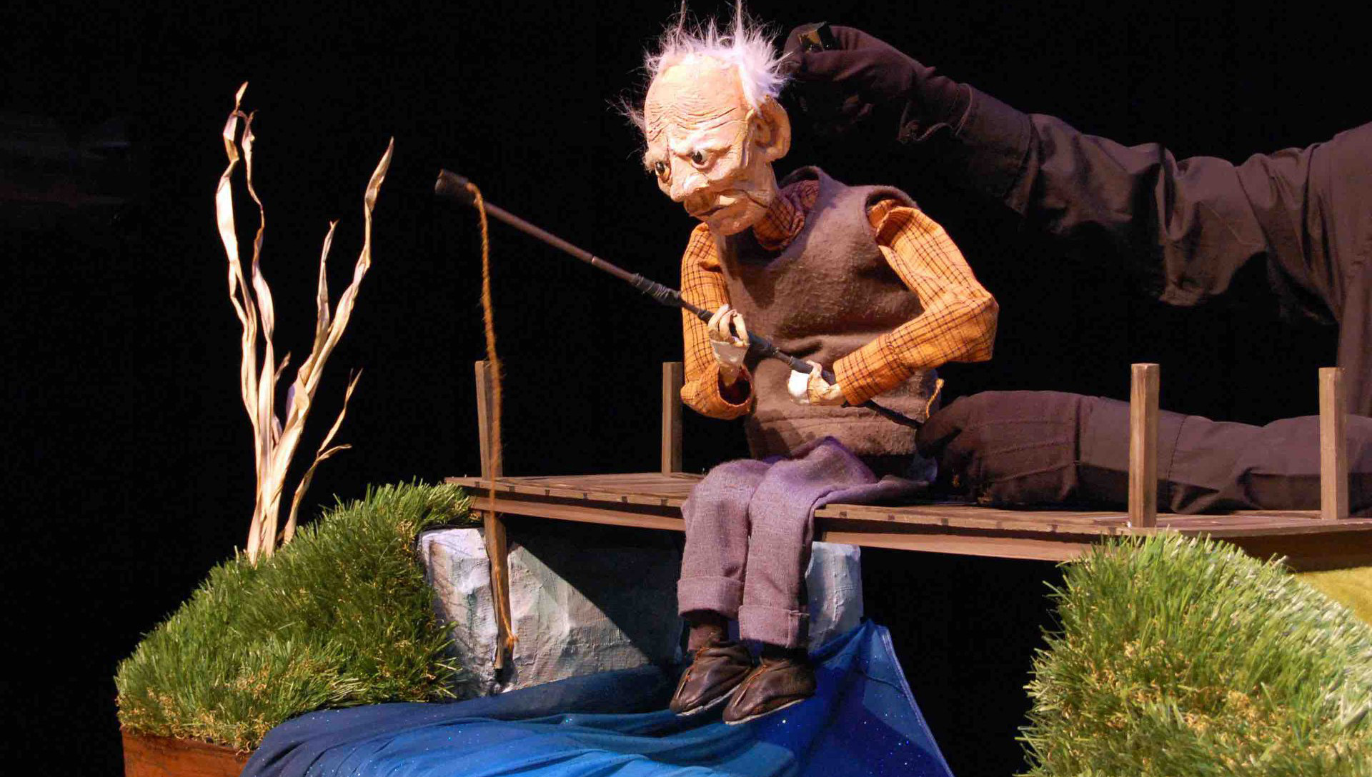 Production shot from the show Old Man and the River. A puppet about 18 inches high of an old man with sparse white hair sits fishing while sitting on a bridge. A river made of blue fabric runs under the bridge and two arms masked with black gloves and sleeves are seen manipulating the puppet.